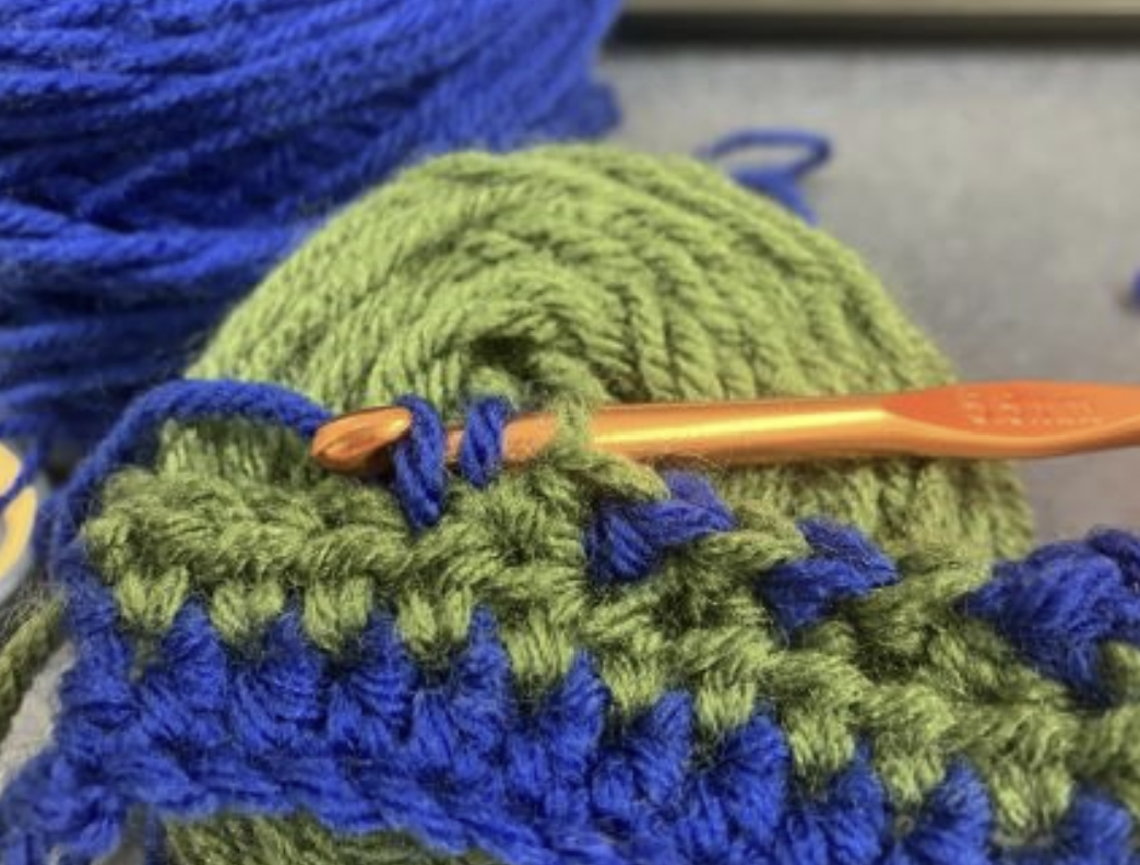 Close-up of a crochet project with green and blue yarn and an orange crochet hook